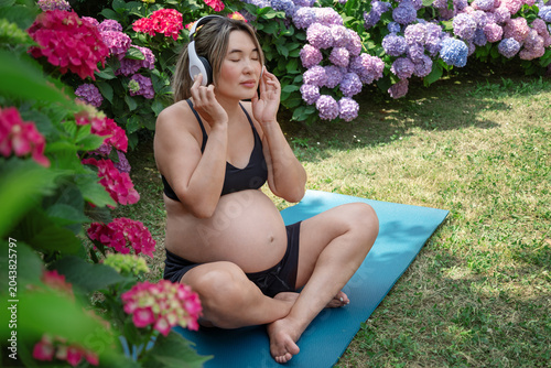 Pregnant asian woman sitting cross legged on a yoga mat in a garden with hydrangeas, listening to music through headphones, her eyes closed in peaceful meditation.Music therapy