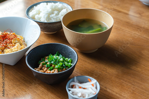 Foto 木製テーブルに並ぶ納豆、紅白なます、味噌汁、ご飯の和食朝食セットの斜め俯瞰写真。ヘルシーな日本の食事の紹介やブログ用