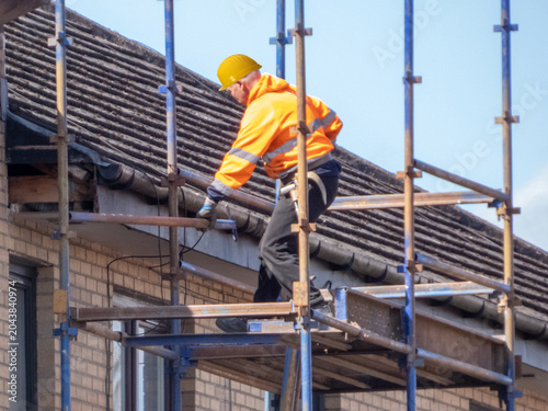 Construction worker is wearing a high-visibility orange jacket and a yellow hard hat.They are performing repairs on a tiled roof while supported by scaffolding. Safe working practices at height. 