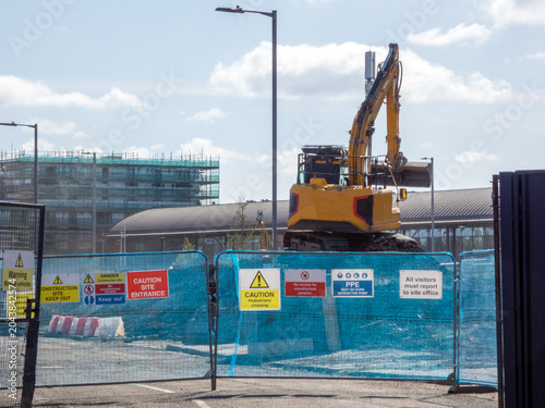A construction site secured by blue fencing panels. Safety signs are displayed, caution notices for site entrances. An excavator is in the background,  in ongoing demolition, and construction work. 