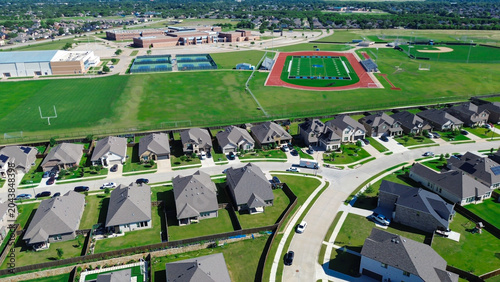Campus linked housing in Arlington, TX shows rows of modern homes, gray roofs, fenced yards parked cars positioned near large school complex. Direct adjacency to athletic fields brick buildings
