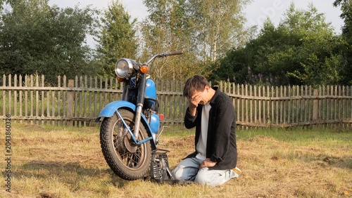 Photography Man kneeling beside a broken blue vintage motorcycle on a rural lawn, holding hi