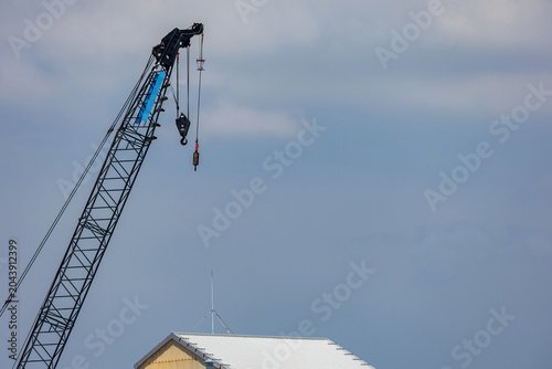 Bild auf Leinwand Diagonally raised lattice crane boom hanging above white roofline of a building set against a pale blue sky, suggesting a pause in work on a construction site