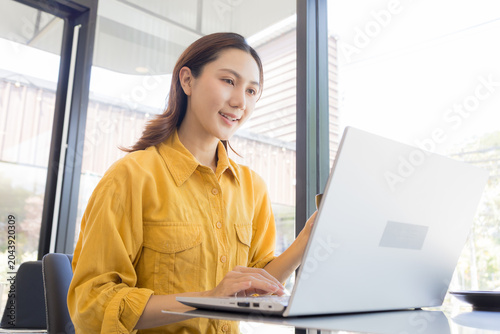 Young woman smiles while holding a coffee cup. She points at her laptop screen, showing something online. Soft daylight shines through a window behind her. Her yellow shirt adds warmth to the scene
