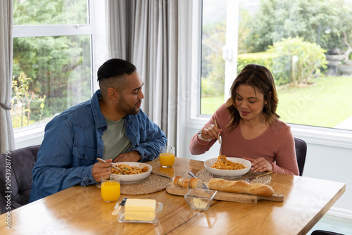 Mid-adult couple sitting at dining table by window twirling spaghetti with baguette and juice