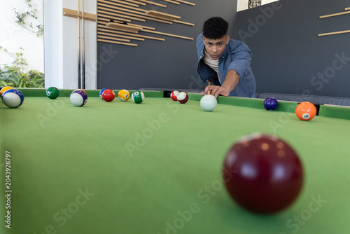 African American teen leaning over green-felt pool table in lounge, aiming cue stick at cue ball