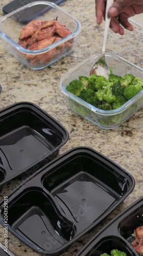 Vertical video: Arm scooping broccoli adding chicken on kitchen counter with empty trays and spoon