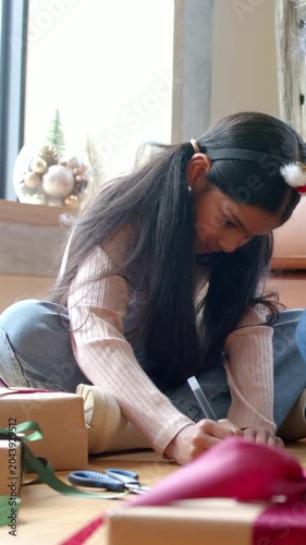 Vertical video: Reaching for pen, Indian schoolgirl in pink top writing gift tag at home