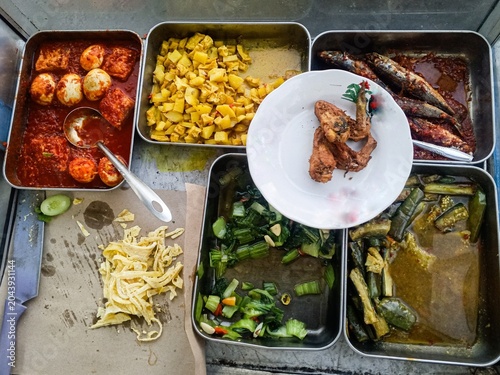 Traditional Indonesian buffet food displayed in stainless steel trays. Various side dishes including eggs in chili sauce, fish, vegetables, tofu, and fried chicken served in a local food stall. Authen