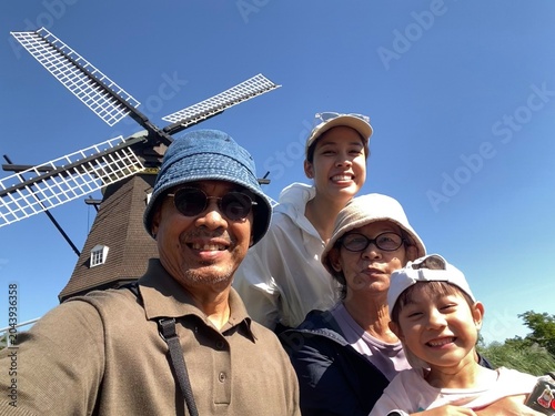 A joyful multi-generational Thai family takes a bright low-angle selfie with a historic European-style windmill under a clear blue sky during an outdoor park excursion in Chiba, Japan.