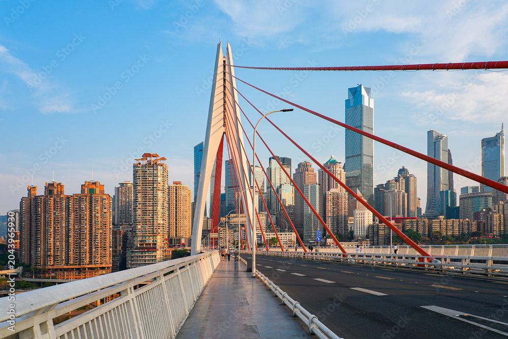 Naklejka premium Pedestrian walkway on Qiansimen Bridge leading to the white pylon and Jiefangbei skyscrapers skyline in Chongqing, China