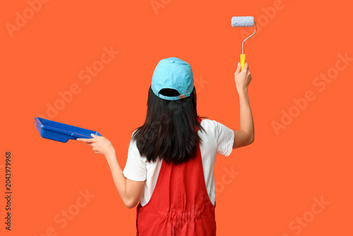 Female Asian painter with roller and tray on orange background, back view