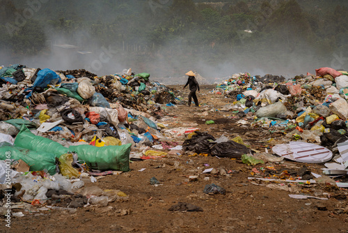 Foto Large piles of mixed garbage accumulate at an open landfill site in Vietnam, with a worker wearing a traditional conical hat walks through a vast landfill surrounded by waste piles and drifting smoke