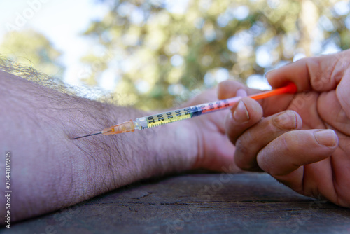 Person's hands using a needle and syringe to inject a solution into their forearm, showing drug addiction or medical treatment outdoors