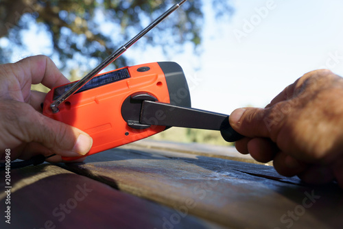 Person winding a portable orange emergency radio, featuring hand crank and solar charging, for survival and outdoor preparedness
