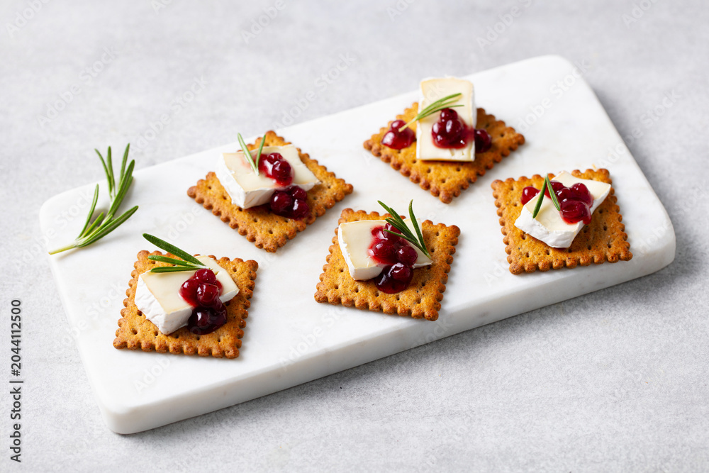 Fototapeta premium Crackers with brie cheese cranberry sauce and fresh rosemary on white marble board. Grey background.