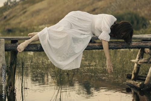 Barefoot woman in a vintage white dress lies on a small wooden bridge over a river and reaching out to the water, magic atmosphere.