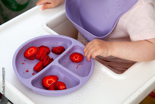 A child sits in a garden on a sunny day, happily eating fresh strawberries. The child has a joyful expression and large, ripe strawberries in hand.