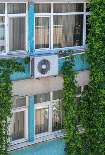 The air conditioner's outdoor unit is on the exterior of the apartment building. Ivy plants covering the exterior of the building.