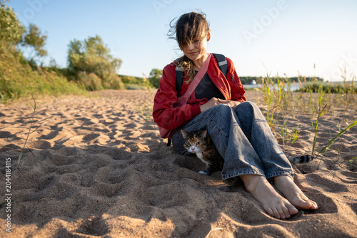 Canvas Print Woman sitting barefoot on wild sandy beach with domestic cat during sunny windy day