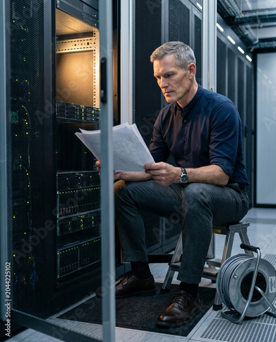 Vertical shot from a slight low angle, focusing on the senior engineer deeply immersed in a multi-page blueprint. Warm tungsten lighting from within the server rack highlights textures.
