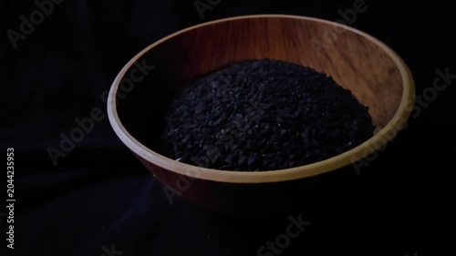 Macro cinematic shot capturing black nigella seeds (Nigella sativa), also known as black caraway, spilling out from a rustic wooden bowl onto a wooden table. The sharp focus and shallow depth of field