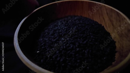 Macro cinematic shot capturing black nigella seeds (Nigella sativa), also known as black caraway, spilling out from a rustic wooden bowl onto a wooden table. The sharp focus and shallow depth of field