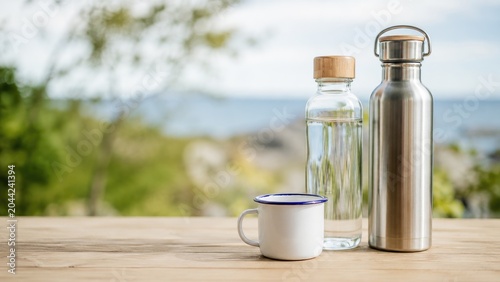 Stainless steel water bottle, glass bottle with wooden lid and white enamel mug on a wooden table by the seaside with a blurred coastal background