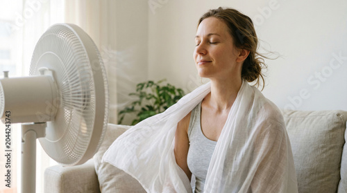 Close up of relaxed young woman with closed eyes enjoying cool air breeze from floor fan on hot summer day at home with flowing transparent fabric