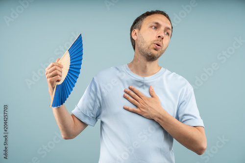 Bild auf Leinwand Middle aged man on blue studio background holding hand fan, sweating, cooling down