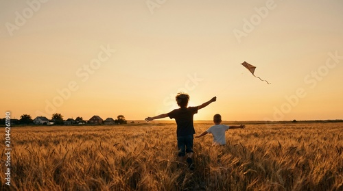 Two boys running with a kite in a wheat field at sunset