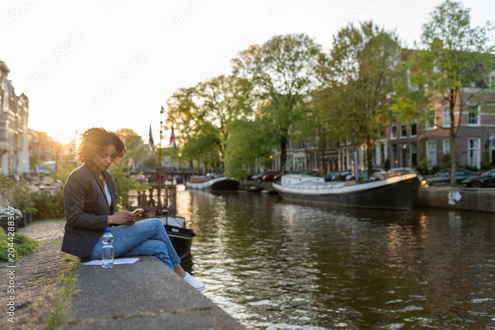 Fototapeta premium Woman freelancer using smartphone while sitting on a canal wall in amsterdam during golden hour