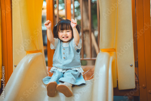 Obraz na plátně Cheerful Asian toddler girl raising her arms in excitement while sliding down a yellow slide at an outdoor playground