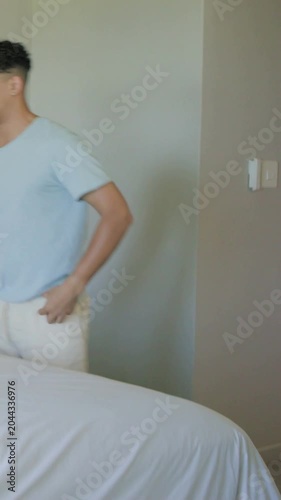 Vertical video: Entering from right, man in blue tee making bed with white sheet in bedroom