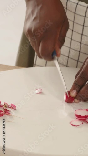 Vertical video: Placing radish, using blue knife, aproned cook hands slicing rounds on white board