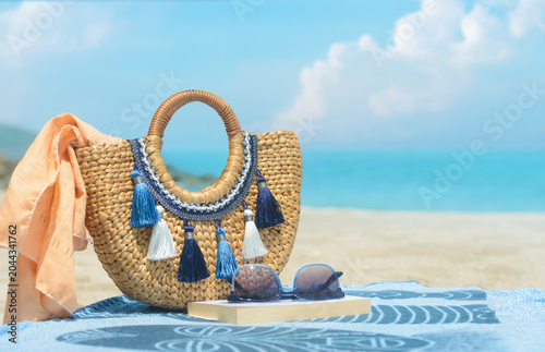 Straw beach bag with tassels, sunglasses and book on towel against blurred sea background. Summer vacation concept