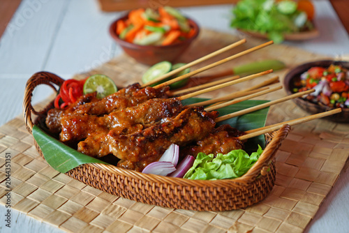 Foto Traditional Indonesian chicken satay with peanut sauce served on bamboo skewers in a woven basket with fresh vegetables and sambal
