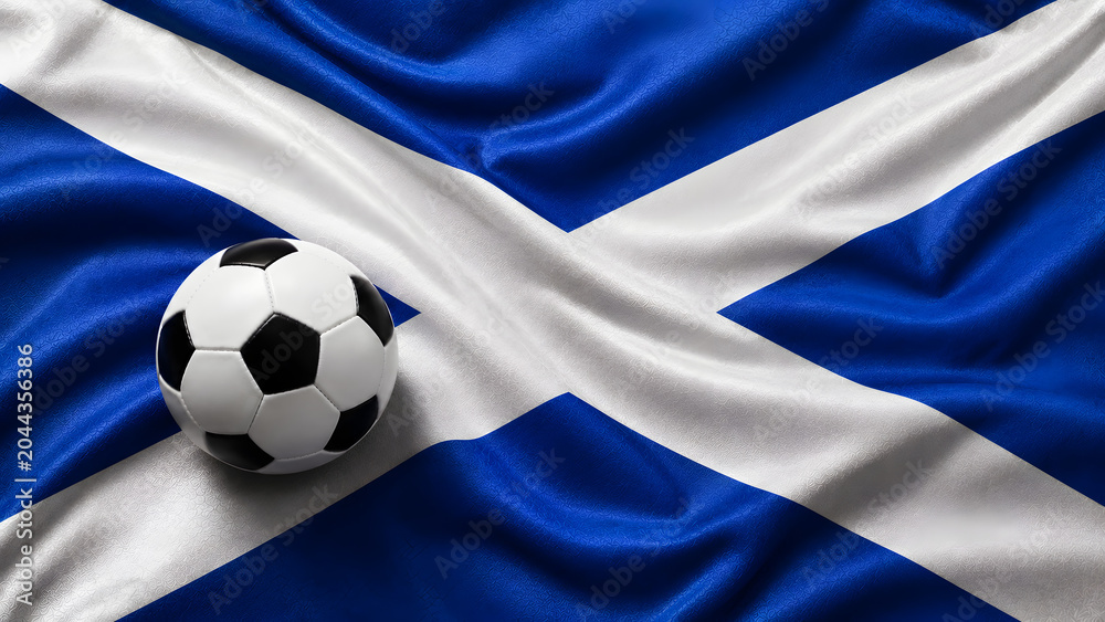 Fototapeta premium A classic black and white soccer ball placed on top of the draped fabric of the Scottish flag, representing football culture and national pride.