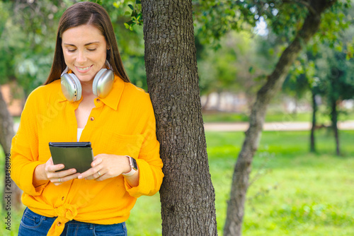 Happy woman wearing yellow shirt holding digital tablet and smiling outdoors in sunny green park with copy space,technology and lifestyle concept, natural background for design or promotional template