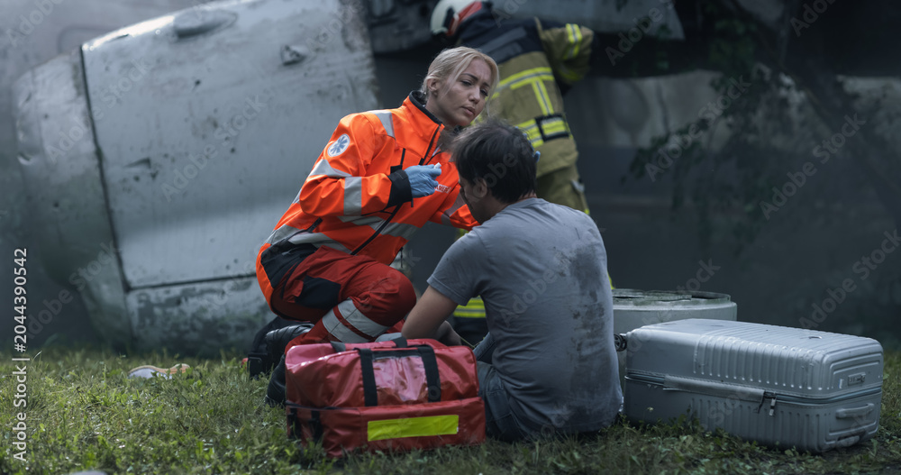 Fototapeta premium A Female Paramedic Evaluates a Seated Male Survivor Next to Damaged Airplane Fuselage Wreckage as a Firefighter Inspects the Crash Site, With Rescue Bag and Luggage Set on the Grass for Emergency Care