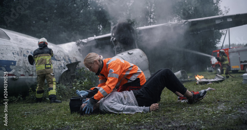 Cuadro en lienzo Female Paramedic Supports an Injured Survivor on the Grass Beside a Wrecked Airplane as Firefighters Cool the Smoking Engine and Fuselage During an Ongoing Coordinated Crash Response Operation
