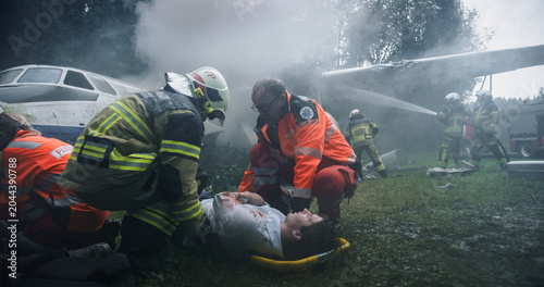 Tableau sur toile Firefighters and Paramedics Work Under a Smoke-filled Airplane Wing at a Forest Crash Site, Lifting Injured Passengers Onto Stretchers While Crews Suppress Fire and Coordinate Emergency Evacuation