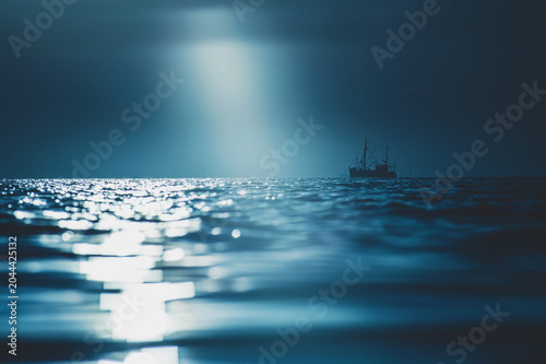 Canvastavla A solitary fishing boat sails on a dark, moonlit ocean with a column of soft light breaking through low cloud and reflecting across rippling water