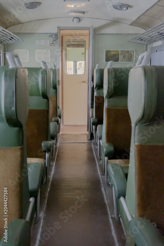 Vintage train carriage interior with central aisle, green seats and worn leather padding. Soft natural light through far door evokes nostalgic rail travel. Empty retro passenger compartment.

