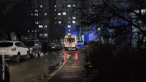 Emergency medical services vehicle van with flashing blue lights parked at entrance of Soviet-style high-rise building. Wet city street at night, residential urban setting