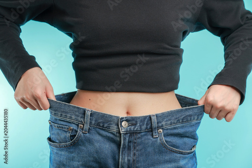 Happy woman showing loose jeans celebrating her healthy weight loss journey with confidence and motivation against bright blue studio background