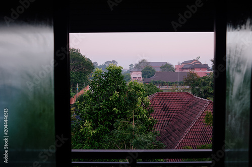 Bilde på lerret Misty morning view through a window over red tile rooftops and tropical trees in a quiet Thai neighborhood