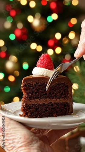 Chocolate cake slice with raspberry topping held on a white plate, fork poised to cut into the dessert, festive Christmas tree with colorful ornaments in the background
