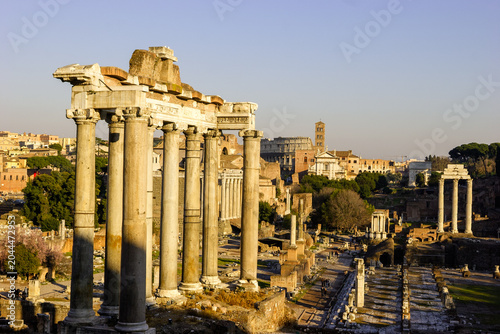 Φωτογραφία The Roman Forum, Rome, Italy, with the Temple of Saturn in the foreground and the Colosseum and Arch of Titus visible in the background under a clear blue sky