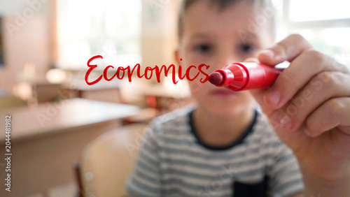 Child with red marker writing economics in school classroom, education learning in elementary setting, young student creativity knowledge concept